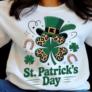 Peut inclure: Sweat-shirt blanc avec un motif de la Saint-Patrick. Le motif comprend un chapeau haut-de-forme vert, un trèfle à quatre feuilles avec un imprimé léopard, des fers à cheval et le texte "St. Patrick's Day" en vert.