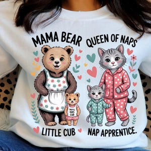 Peut inclure: Sweat-shirt blanc avec un motif de famille ours et chat. L'ours porte un tablier, le chat porte un pyjama. Le texte dit "Mama Bear Queen of Naps Little Cub Nap Apprentice."