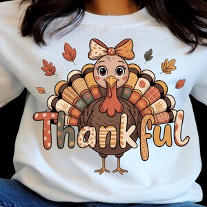 Peut inclure: Sweat-shirt blanc avec un dessin de dinde de dessin animé. La dinde porte un nœud, des plumes de queue à motifs et le mot "Thankful". Le motif comprend des feuilles d'automne et des motifs bruns, oranges et beiges.