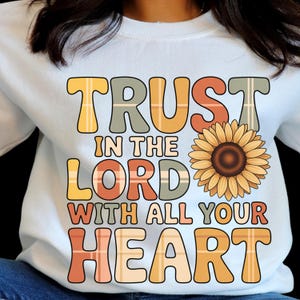 Peut inclure: Sweat-shirt blanc avec l'inscription "TRUST IN THE LORD WITH ALL YOUR HEART" dans un design à carreaux coloré. Un grand graphisme de tournesol est à côté du texte. Le sweat-shirt est de style décontracté et confortable.
