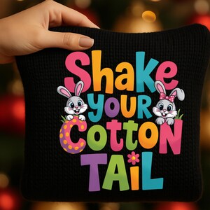 Peut inclure: Coussin noir avec le texte "Shake Your Cotton Tail" en lettres colorées. Le motif comprend deux lapins de dessin animé et une fleur. Le coussin est tenu par une personne.