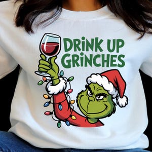 Peut inclure: Sweat-shirt blanc avec un motif du Grinch tenant un verre de vin rouge. Le Grinch porte un bonnet de Père Noël et un manteau rouge. Le texte "DRINK UP GRINCHES" est en vert. Vêtements de Noël.