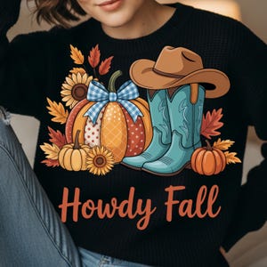 Peut inclure: Pull noir avec un motif sur le thème de l'automne. Le motif représente une citrouille avec un nœud vichy bleu, des tournesols, des feuilles d'automne, des bottes de cowboy et un chapeau de cowboy. Les mots "Howdy Fall" sont écrits en orange.