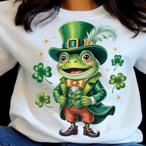 Peut inclure: Sweat-shirt blanc avec une grenouille de dessin animé habillée en leprechaun. La grenouille porte un haut-de-forme vert, un manteau et un nœud papillon, avec un pantalon marron et des chaussures noires. Des trèfles verts et des étoiles dorées décorent le motif.