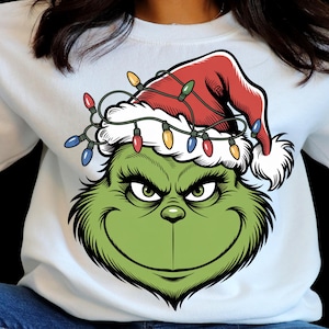May include: White sweatshirt featuring a green cartoon character face with a red Santa hat and string lights. The character has a mischievous grin and is a popular holiday icon. The sweatshirt is a festive apparel item.