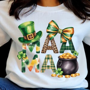 Pode incluir: Camisola branca com um design do Dia de São Patrício. A palavra "MAMA" é exibida com um chapéu de duende verde, um laço e um pote de ouro. As letras têm vários padrões, incluindo xadrez e estampa de leopardo, com trevos.
