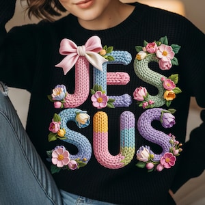 Peut inclure: Pull-over noir avec le mot "JESUS" en lettres colorées et texturées. Les lettres sont ornées d'éléments floraux et d'un nœud rose. Le pull est un vêtement confortable et élégant.