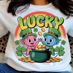 Peut inclure: Sweat-shirt blanc avec un motif coloré représentant deux lutins de dessin animé, un pot d'or et un arc-en-ciel. Le mot "LUCKY" est écrit au-dessus des lutins en grandes lettres colorées. Le motif comprend des trèfles et des pièces d'or.