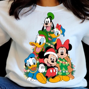 May include: White sweatshirt featuring a Christmas tree design made of Disney characters, including Mickey Mouse, Minnie Mouse, Donald Duck, Daisy Duck, and Goofy. The characters are in festive attire, with Mickey wearing a Santa hat.