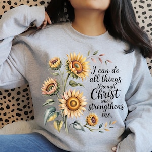 Puede incluir: Una sudadera gris claro con un diseño de acuarela de girasoles en tonos amarillos, naranjas y verdes. El texto "I can do all things through Christ who strengthens me" está impreso en la parte delantera.