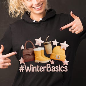 Peut inclure: Un sweat à capuche noir avec un motif d'accessoires d'hiver : un sac à main marron, des bottes jaunes, une tasse à café et un bonnet jaune. Le texte "#WinterBasics" est affiché en dessous.