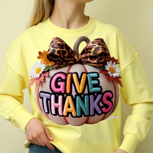 Peut inclure: Un sweat-shirt jaune avec un grand motif de citrouille décorative. La citrouille beige porte l'inscription "GIVE THANKS" en lettres brodées colorées. Un nœud léopard et des décorations florales ornent le dessus de la citrouille, créant un thème automnal festif.