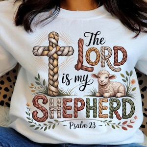 Peut inclure: Sweat-shirt blanc avec une croix tressée et l'inscription "The Lord is my Shepherd" avec une illustration d'agneau. Le motif comprend le texte "Psaume 23" en bas, avec des accents floraux. Le sweat-shirt est de style décontracté et confortable.