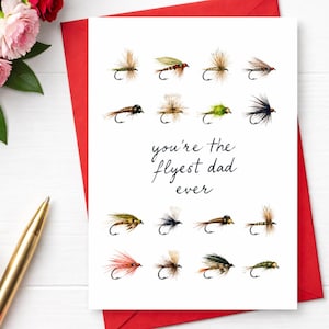 Puede incluir: Una tarjeta blanca con un sobre rojo presenta ilustraciones de acuarela de moscas de pesca. El texto en cursiva dice "you're the flyest dad ever". La tarjeta está rodeada de flores y un bolígrafo dorado.