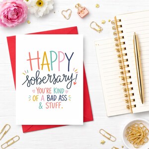May include: A white greeting card with the words "HAPPY sober-sary!" in colorful lettering, inside a red envelope. The card also says "YOU'RE KIND OF A BAD ASS & STUFF." A gold pen, notepad, and other stationery items are arranged around the card.
