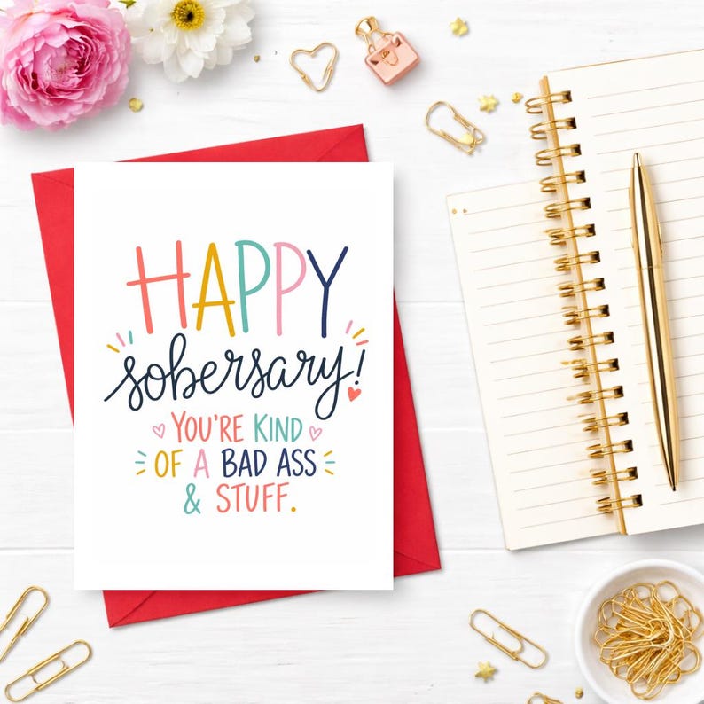 May include: A white greeting card with the words "HAPPY sober-sary!" in colorful lettering, inside a red envelope. The card also says "YOU'RE KIND OF A BAD ASS & STUFF." A gold pen, notepad, and other stationery items are arranged around the card.