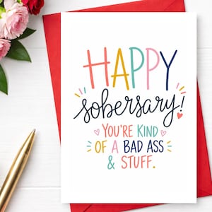 May include: A white greeting card with the words "HAPPY sobersary!" in colorful lettering. The card also includes the phrases "YOU'RE KIND of A BAD ASS & STUFF." A red envelope and a gold pen are visible.