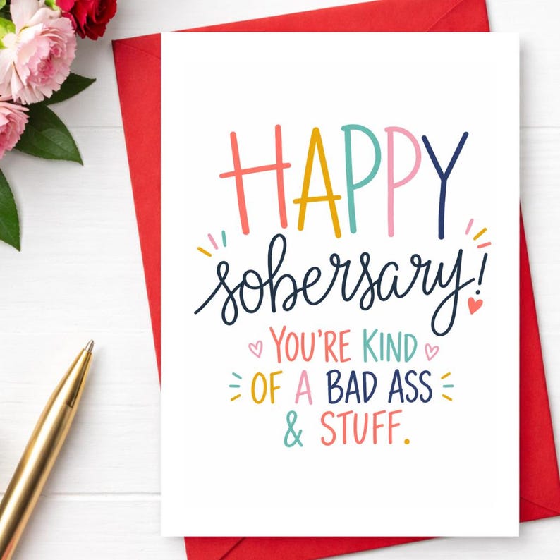May include: A white greeting card with the words "HAPPY sobersary!" in colorful lettering. The card also includes the phrases "YOU'RE KIND of A BAD ASS & STUFF." A red envelope and a gold pen are visible.