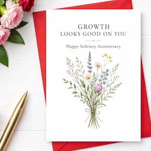 May include: A white greeting card featuring a watercolor illustration of a wildflower bouquet tied with twine. The text reads: GROWTH LOOKS GOOD ON YOU. Happy Sobriety Anniversary. The card is positioned on a red envelope with a gold pen nearby.