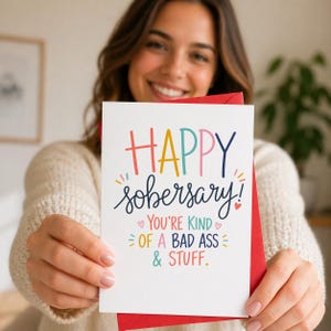 May include: A white greeting card with the words "HAPPY sobersary!" in colorful lettering. The card also includes the text "YOU'RE KIND OF A BAD ASS & STUFF." The card is held in front of a red envelope.