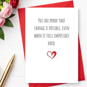 Puede incluir: Una tarjeta de felicitación blanca con el texto "YOU ARE PROOF THAT CHANGE IS POSSIBLE, EVEN WHEN IT FEELS IMPOSSIBLY HARD." Un corazón rojo en la parte inferior. La tarjeta está dentro de un sobre rojo.