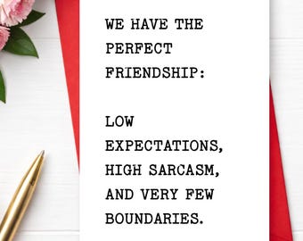 May include: A white greeting card with black typewriter-style text that reads: WE HAVE THE PERFECT FRIENDSHIP: LOW EXPECTATIONS, HIGH SARCASM, AND VERY FEW BOUNDARIES. The card is placed on a red envelope, next to a gold pen and pink flowers.