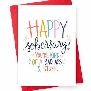 May include: A white greeting card with a red envelope. The card has the words "HAPPY sobersary!" in colorful letters. Below, it reads "YOU'RE KIND OF A BAD ASS & STUFF." Small hearts and decorative lines are also included.