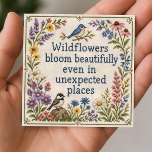 May include: Cross-stitch square with the text "Wildflowers bloom beautifully even in unexpected places." The design features a border of colorful flowers and two birds. The color palette includes blues, purples, yellows, and greens.