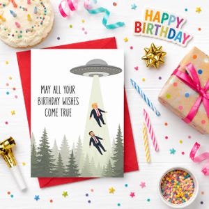 May include: A rectangular birthday greeting card featuring a gray UFO hovering over a forest, beaming down two figures in suits. The text reads, "MAY ALL YOUR BIRTHDAY WISHES COME TRUE." The card is set against a white background with confetti.