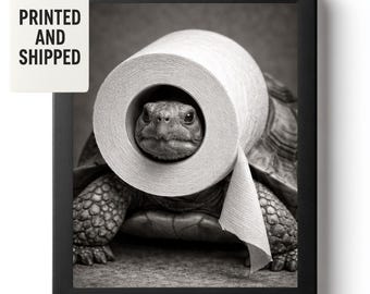 Könnte beinhalten: Schwarz-Weiß-Druck in einem Rahmen, der eine Schildkröte mit einer Toilettenpapierrolle um den Kopf zeigt. Die Schildkröte steht im Mittelpunkt, wobei die Rolle ihr Gesicht umrahmt. Der Druck trägt den Text "PRINTED AND SHIPPED" auf einem Anhänger.