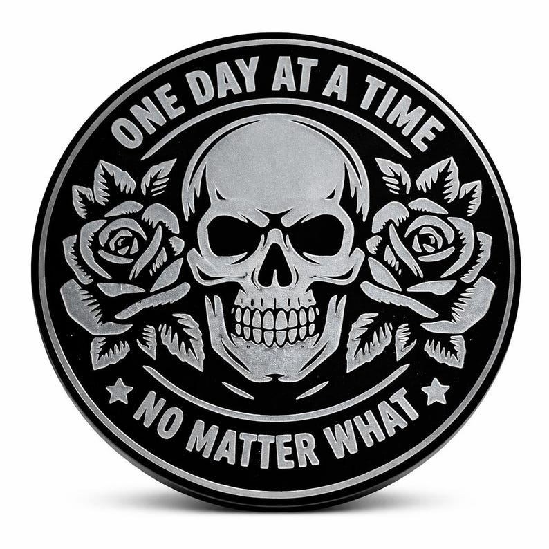 May include: A black and silver round emblem featuring a skull, roses, and the phrases "ONE DAY AT A TIME" and "NO MATTER WHAT". The skull is in the center, with roses on either side.