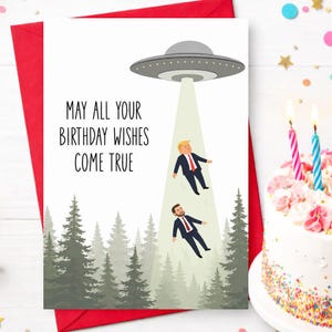 May include: A white birthday greeting card featuring a gray UFO hovering over a forest of green pine trees. A light beam shines down, lifting two figures in dark suits and red ties. The text reads, May all your birthday wishes come true.