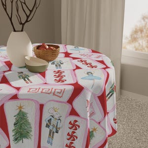 May include: A round red tablecloth with a repeating pattern of white rectangles featuring watercolor illustrations of a Christmas tree, a mouse soldier, a ballerina, and other holiday-themed images. A vase, bowl, and basket are on the table.