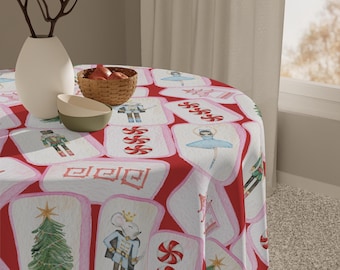 Op de afbeelding: Een ronde rode tafelkleed met een herhalend patroon van witte rechthoeken met aquarel illustraties van een kerstboom, een muizensoldaat, een ballerina en andere afbeeldingen met een vakantiethema. Een vaas, een kom en een mand staan op de tafel.