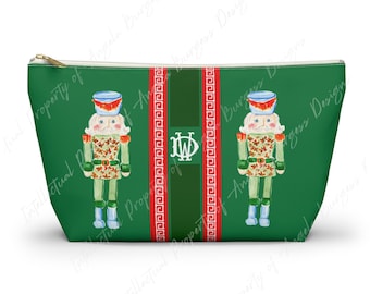 May include: A green zippered pouch featuring two illustrated nutcracker figures with red and white accents. A central vertical stripe contains a white monogram with the letters DW, flanked by two red borders with a white Greek key pattern.