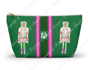 May include: A green zippered pouch featuring two illustrated nutcrackers in pink and green uniforms. A central vertical dark green stripe is flanked by two bright pink bands with a white Greek key pattern and a white monogram with the letters DW.