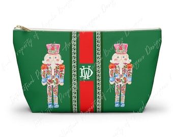 May include: A green zippered pouch featuring two nutcracker illustrations with floral patterns, pink hats, and blue accents. A central vertical red stripe is flanked by white Greek key borders and contains a white monogram with the letters DW.