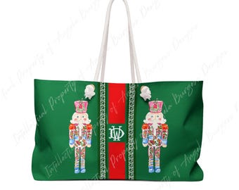 May include: A green tote bag with white rope handles featuring two illustrated nutcrackers with floral patterns. A central vertical red stripe contains a white monogram with the letters JW. The design includes white Greek key borders.