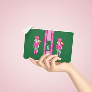 May include: A green rectangular zippered pouch featuring two pink nutcracker figures holding lollipops on either side of a central vertical pink stripe. The stripe contains the white monogram letters CN in a decorative serif font with a Greek key border.