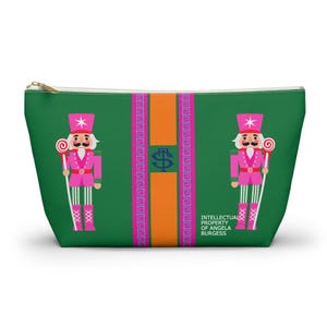 May include: Green zippered pouch featuring a pink and white nutcracker soldier design. The pouch has a gold zipper and a central vertical stripe in orange and pink. Text on the right side reads "INTELLECTUAL PROPERTY OF ANGELA BURGESS."