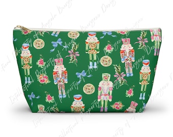 May include: A green zippered pouch featuring a repeating pattern of nutcracker figures, floral bouquets, and light blue bows. The nutcrackers are depicted in various poses with colorful uniforms, and the design includes small circular floral motifs.