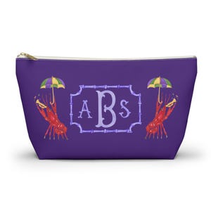 May include: A purple zippered pouch featuring a decorative design. The design includes a bamboo-style frame with the letters "ABS" inside. Two red crawfish with umbrellas flank the frame. The pouch has a gold zipper.
