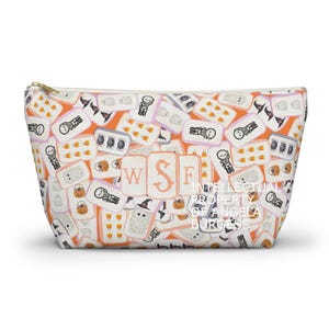 May include: A zippered cosmetic bag with a Halloween-themed pattern. The design features various Halloween icons like ghosts, candy corn, and skeletons on an orange background. The bag has the text "WSF INTELLECTUAL PROPERTY OF ANGELA BURGESS".