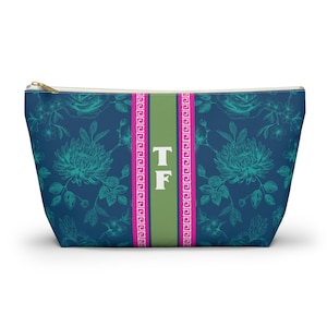 May include: A navy blue zippered pouch featuring a teal floral pattern and a central vertical olive green stripe. The stripe is bordered by two bright pink bands with a white Greek key design and displays the white capital letters TF.