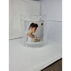 May include: A clear glass picture frame with a curved front and a molded leaf pattern along the border. The frame contains a photograph of a person wearing a white veil and dress holding a bouquet, with the text Princess House printed at the bottom.