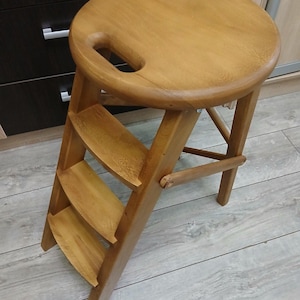 Op de afbeelding: Een houten opstapje met een ronde zitting en een ingebouwde handgreep. De kruk heeft drie treden en is gemaakt van lichtgekleurd hout. Het ontwerp is functioneel en ruimtebesparend, geschikt voor het bereiken van hoge planken of kasten.