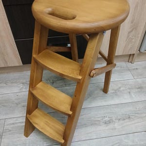 May include: A wooden step stool with a circular seat and three steps. The stool is made of light-coloured wood and has a handle cut into the seat. The steps fold out for use and fold in for storage.