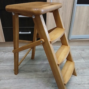 Peut inclure: Un escabeau en bois avec un dessus rectangulaire et trois marches. L'escabeau est en bois de couleur claire et a un design pliable. L'escabeau mesure environ 46 cm de haut.