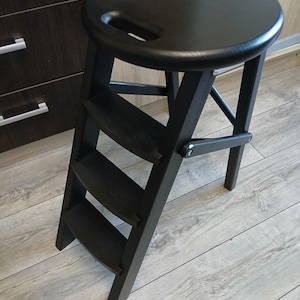 Könnte beinhalten: Ein schwarzer Holztritt mit rundem Sitz und integriertem Griff. Der Hocker hat drei Stufen und ein Klappdesign. Der Hocker ist aus dunklem Holz gefertigt und für das Erreichen höherer Stellen konzipiert.
