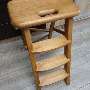 May include: A wooden step stool with a light brown finish. The stool has a rectangular top with a handle cutout and three curved steps. The design is simple and functional, suitable for reaching higher places.